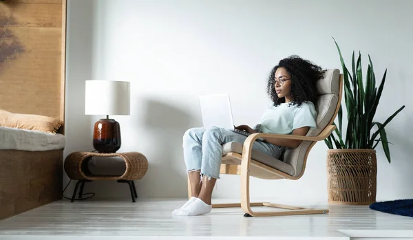 Όμορφη νεαρή χαμογελαστή Αφροαμερικανή γυναίκα που εργάζεται σε φορητό υπολογιστή, ενώ κάθεται σε καρέκλα άνετα στο σπίτι — Φωτογραφία Αρχείου