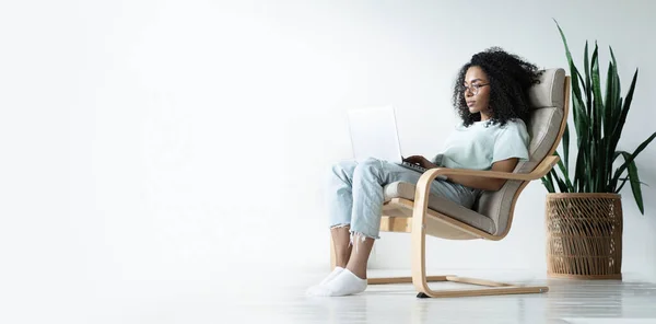 Belle jeune femme afro-américaine souriante qui travaille sur un ordinateur portable tout en étant assise dans une chaise confortable à la maison — Photo