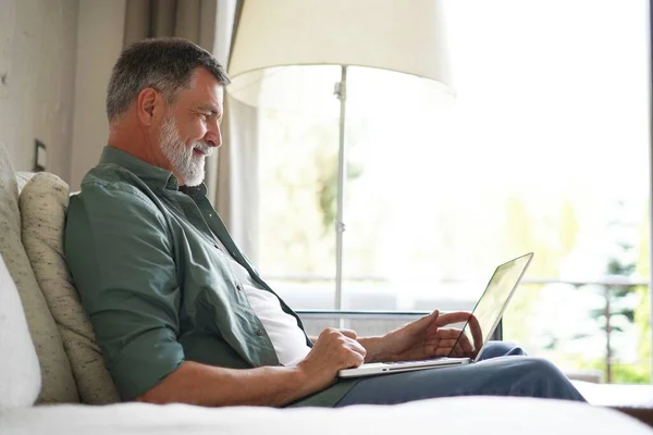 Retrato de homem maduro feliz em roupas casuais usando laptop deitado no sofá em casa. — Fotografia de Stock