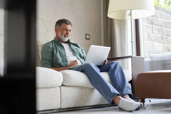 Portré boldog érett férfi alkalmi ruhák segítségével laptop fekvő kanapén a házban. — Stock Fotó