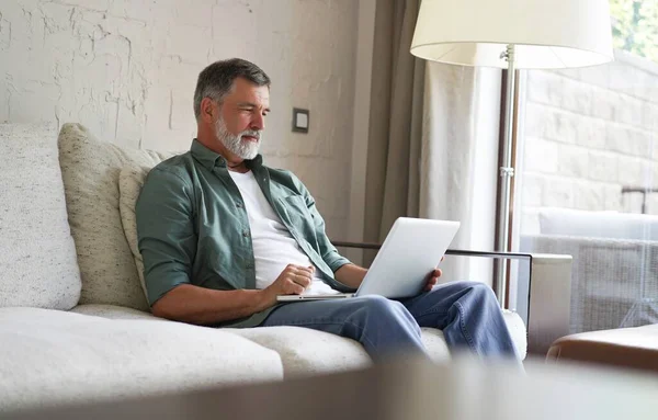 Retrato de homem maduro feliz em roupas casuais usando laptop deitado no sofá em casa. — Fotografia de Stock