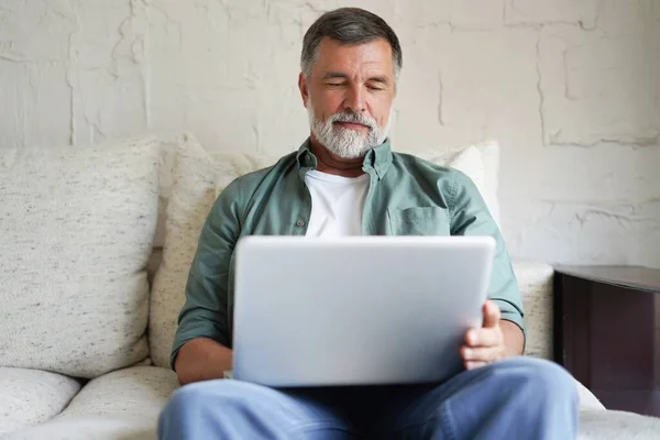 Retrato de homem maduro feliz em roupas casuais usando laptop deitado no sofá em casa. — Fotografia de Stock