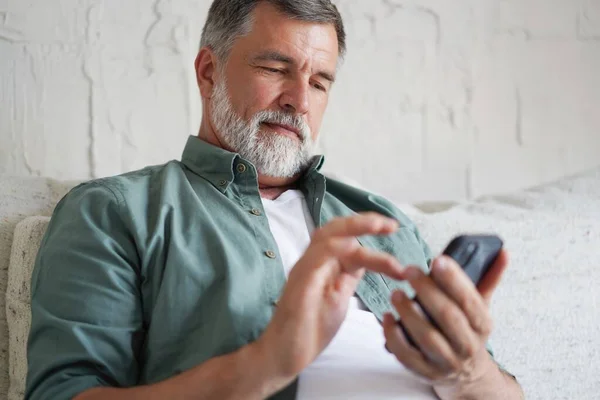 Sorrindo Homem Sênior Usando Celular Navegação Internet Sentado no Sofá Interior. — Fotografia de Stock