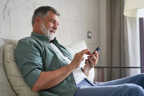 Mosolyogva Senior Man használata mobiltelefon böngészés Internet Ülés a kanapén beltéri. — Stock Fotó