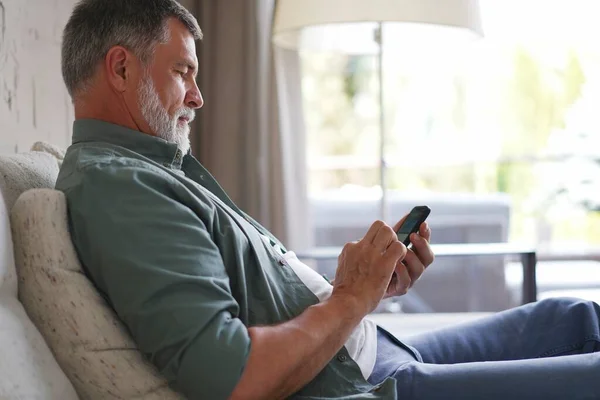 Hombre mayor sonriente usando el teléfono celular navegando por Internet sentado en el sofá interior. — Foto de Stock