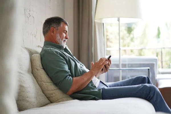 Sorrindo Homem Sênior Usando Celular Navegação Internet Sentado no Sofá Interior. — Fotografia de Stock