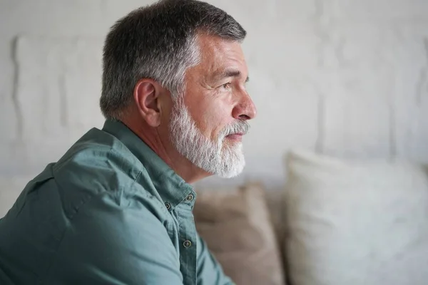 Portrait of attractive pensive senior man sitting in sofa at home and looking at window. — Stockfoto