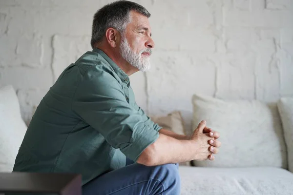 Portrait of attractive pensive senior man sitting in sofa at home and looking at window. — Stockfoto