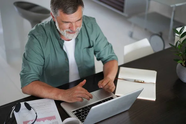 Knappe volwassen man in casual pak zitten aan de tafel in huis kantoor en werken op laptop — Stockfoto