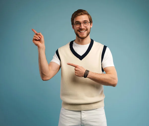 Portrait of a happy young man in casual pointing fingers away at copy space isolated over blue background. — Stock Photo, Image