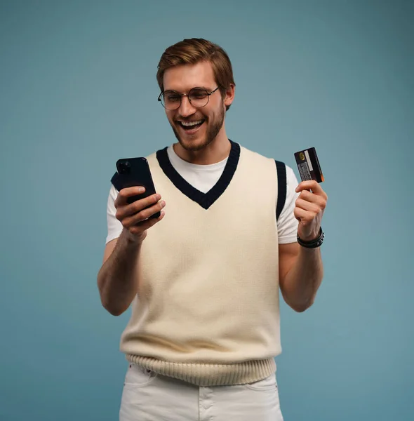 Porträt eines aufgeregten jungen Mannes, der sein Handy in der Hand hält und seine Kreditkarte isoliert vor blauem Hintergrund zeigt. — Stockfoto