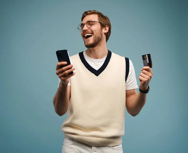 Porträt eines aufgeregten jungen Mannes, der sein Handy in der Hand hält und seine Kreditkarte isoliert vor blauem Hintergrund zeigt. — Stockfoto