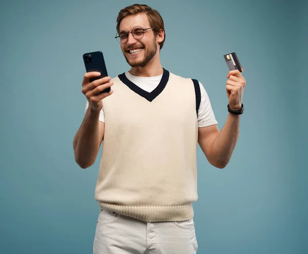 Porträt eines aufgeregten jungen Mannes, der sein Handy in der Hand hält und seine Kreditkarte isoliert vor blauem Hintergrund zeigt. — Stockfoto