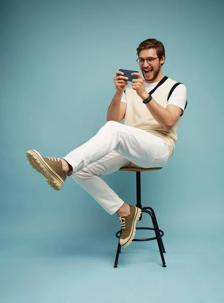 Porträt eines aufgeregten jungen Mannes, der auf einem Schemel sitzt und auf blauem Hintergrund isoliert mit dem Handy spielt. — Stockfoto