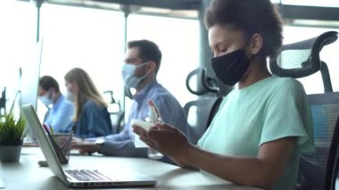 Tıbbi maske takan melez bir kadın ofis işyerinde oturan bilgisayar klavyesini temizliyor.