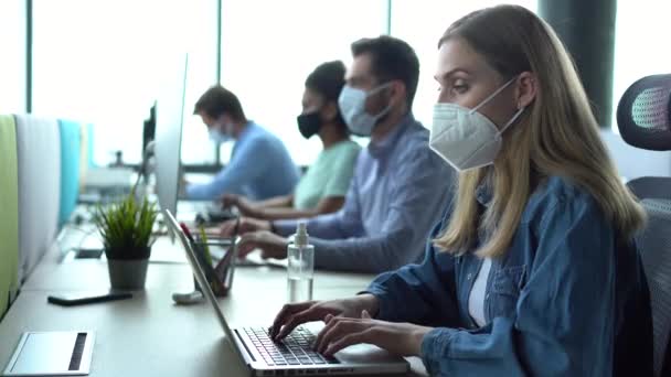 Tıbbi maske takan bir kadın ofis işyerinde oturan bilgisayar klavyesini temizliyor. — Stok video