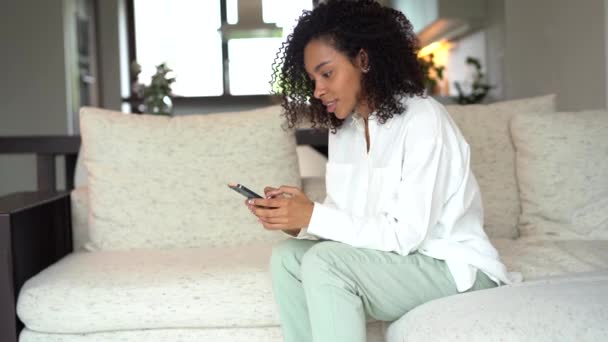 Happy young mixed race woman relaxing on couch, using smartphone, typing message — 图库视频影像