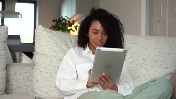 Молодая женщина смешанной расы сидит на диване, используя цифровой компьютер планшет дома — стоковое видео