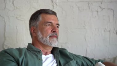 Kanepede dinlenen gülümseyen olgun bir adam. Gri saçlı, evde dinlenen ve kameraya bakan kendinden emin son sınıf öğrencisi.
