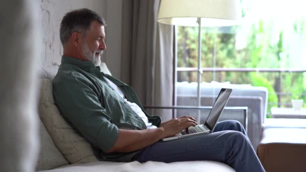 Mayores de edad madura hombre escribiendo en el ordenador portátil de navegación por Internet utilizando aplicaciones sentarse en el sofá solo — Vídeos de Stock