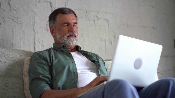 Mayores de edad madura hombre escribiendo en el ordenador portátil de navegación por Internet utilizando aplicaciones sentarse en el sofá solo — Vídeos de Stock