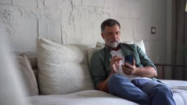 Gülümseyen yaşlı gri saçlı adam koltukta oturuyor ve cep telefonu kullanıyor, sohbet ediyor ya da mobil uygulama kullanıyor.