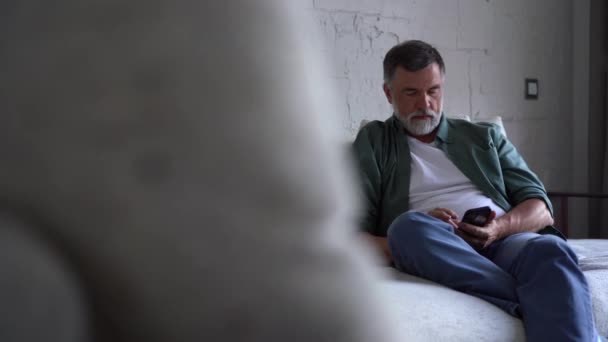Leende äldre gråhårig man som sitter på soffan och använder mobiltelefon hemma, pratar eller använder mobilapplikation — Stockvideo