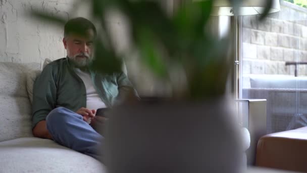 Hombre mayor de pelo gris sonriente sentado en el sofá y usando el teléfono móvil en casa, charlando o usando la aplicación móvil — Vídeo de stock
