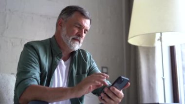Gülümseyen yaşlı gri saçlı adam koltukta oturuyor ve cep telefonu kullanıyor, sohbet ediyor ya da mobil uygulama kullanıyor.
