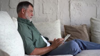 Gülümseyen gri saçlı yetişkin adam internette tabletle sörf yapıyor.