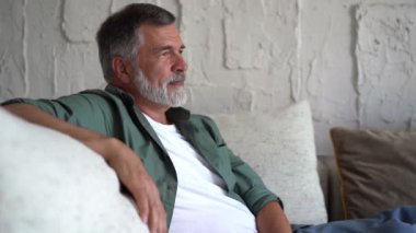Kanepede dinlenen gülümseyen olgun bir adam. Gri saçlı, evde dinlenen ve kameraya bakan kendinden emin son sınıf öğrencisi.