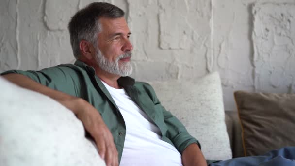 Lachende volwassen man ontspannen op de bank. Zelfverzekerde oudere man met grijs haar die thuis rust en naar de camera kijkt — Stockvideo