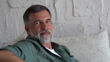 Kanepede dinlenen gülümseyen olgun bir adam. Gri saçlı, evde dinlenen ve kameraya bakan kendinden emin son sınıf öğrencisi.