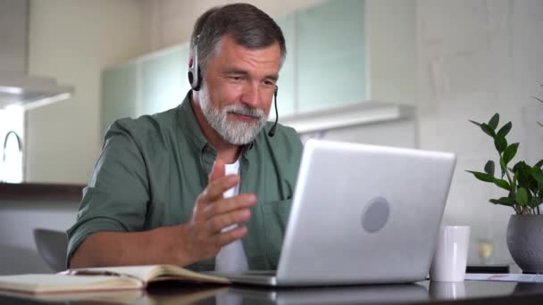 Senior grijsharige man draagt draadloze koptelefoon waardoor videoconferentie zakelijke oproep op laptop zitten thuis bureau — Stockvideo
