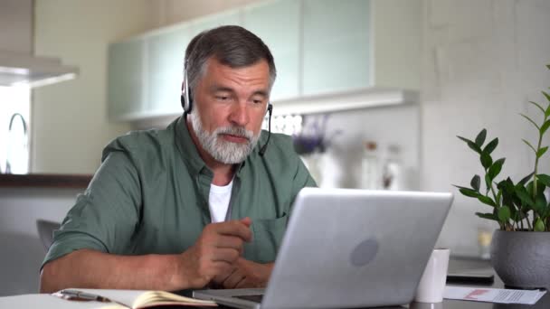 Senior grauhaarige Mann tragen drahtlose Kopfhörer machen Videokonferenz Business-Anruf auf Laptop sitzen zu Hause im Büro Schreibtisch