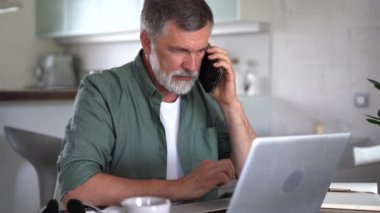Yakışıklı, ciddi, olgun bir adam evde dizüstü bilgisayarla çalışırken telefonla konuşuyor.
