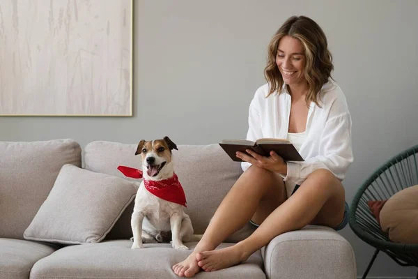 Mujer joven leyendo libro y su lindo perro en el sofá en casa. Hermosa mascota. — Foto de Stock