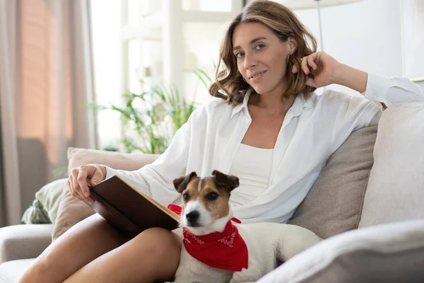 Jovem mulher lendo livro e seu cão bonito no sofá em casa. Bonito animal de estimação. — Fotografia de Stock