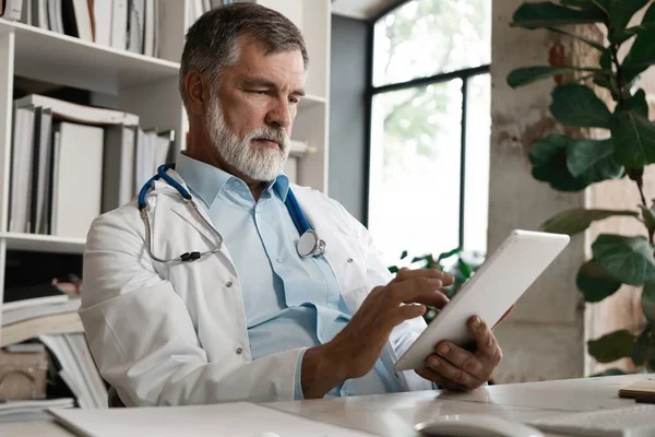 Vrolijke volwassen arts met behulp van digitale tablet in kliniek. — Stockfoto