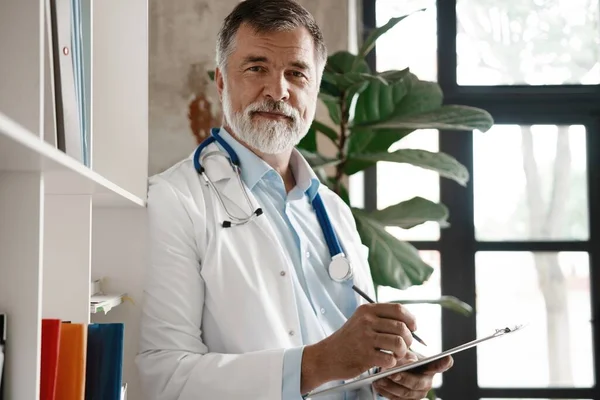 Porträt eines gut aussehenden reifen Arztes, der im modernen Büro am Fenster steht. — Stockfoto