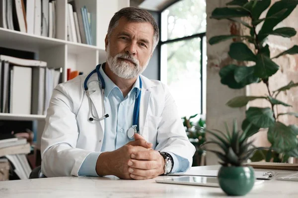 Retrato de belo médico maduro sentado na mesa no escritório moderno. Imagem De Stock