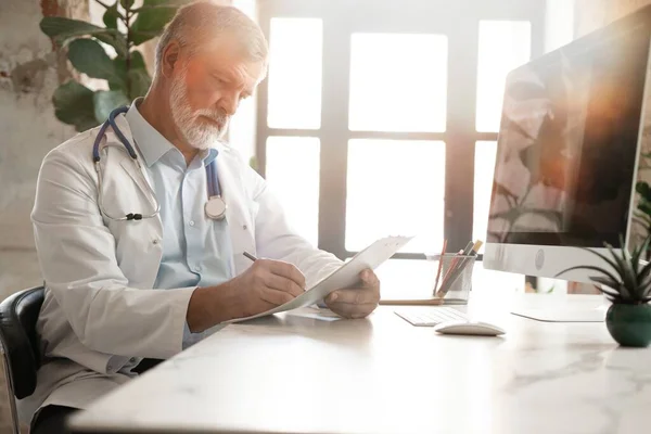 잘생긴 의사가 책상에 앉아서 환자의 의료 카드에 메모를 하거나 작성하거나 약을 처방하는 모습 — 스톡 사진