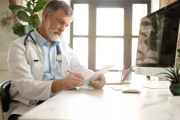 Az asztalnál ülő jóképű orvos portréja, aki jegyzetel vagy kitölti az ügyfelek orvosi kártyáját, vagy gyógyszert ír fel — Stock Fotó