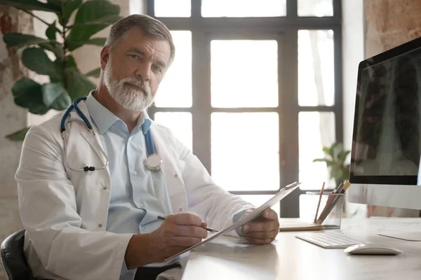 Az asztalnál ülő jóképű orvos portréja, aki jegyzetel vagy kitölti az ügyfelek orvosi kártyáját, vagy gyógyszert ír fel — Stock Fotó