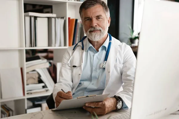 Retrato de médico bonito sentado na mesa, tomando notas ou preenche os clientes cartão médico ou prescreve medicação — Fotografia de Stock