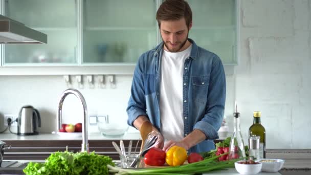 Genç ve mutlu bir adam evde tek başına sağlıklı yiyecekler hazırlıyor. Modern mutfak içinde taze sebze salatası kesiyor. — Stok video