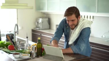 Tablette tarifi takip eden ve evde lezzetli yemekler pişiren bir adam.