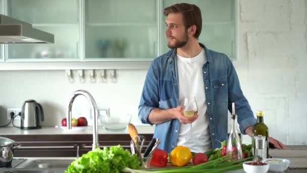 Relaxado jovem bebendo vinho perto da mesa com ingredientes frescos na cozinha — Vídeo de Stock