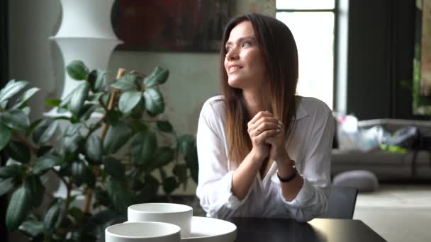 Joven atractiva mujer caucásica vistiendo ropa casual mirando a la cámara y sonriendo se sienta en un café — Vídeos de Stock