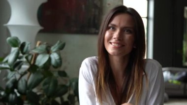 Gündelik giysiler giymiş, kameraya bakıp gülümseyen beyaz bir kadın bir kafede oturuyor.